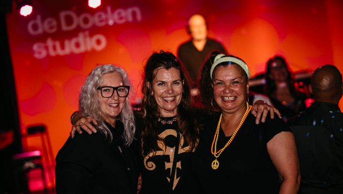 Three smiling women pose together for the camera during Disco Royale at de Doelen Studio. They stand close together in a festive atmosphere. The background is lit in red, with on the wall the words: de Doelen studio. Behind them, a DJ stands on the stage.