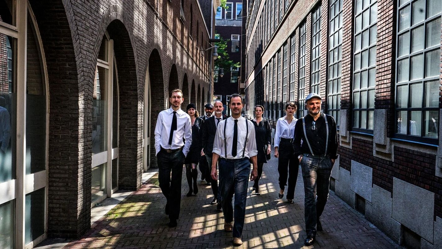Group photo of six men dressed in dark suits with white shirts, standing in a straight line against a concrete wall. They look serious and professional toward the camera.