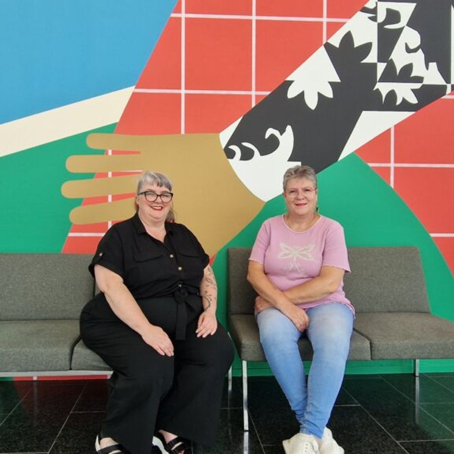 Twee vrouwen zitten glimlachend op een bank voor een kleurrijke muur met grafische vormen en een gestileerde handdruk als achtergrond.