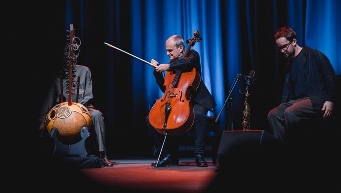 Vier muzikanten spelen in een intieme, blauw verlichte setting op accordeon, kora, cello en een klein snaarinstrument tijdens een sfeervol podiumoptreden.