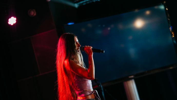 Een jonge zangeres staat solo op het podium tijdens de Vocal Night van de Jordan Roy Vocal Academy in Studio 1 van de Doelen. Ze zingt vol overgave onder warme theaterlichten, tegen een donkere achtergrond.