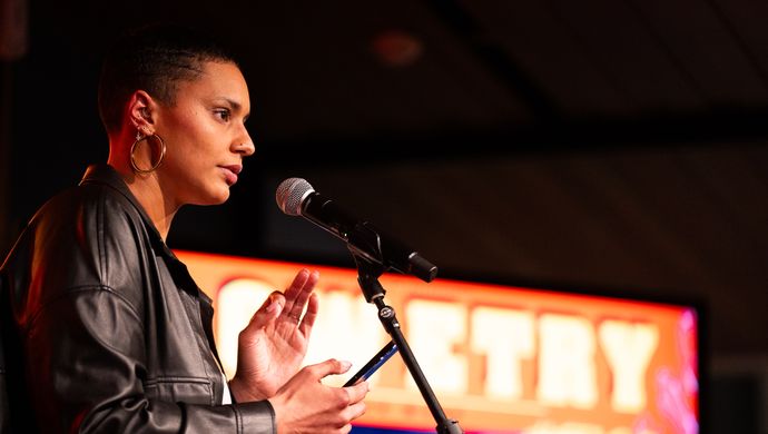 Vrouw met kort haar spreekt in een microfoon op een podium, gekleed in een leren jasje. Op de achtergrond is een kleurrijk scherm te zien met het woord “POETRY”.