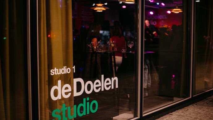 Exterior view of the glass facade of de Doelen Studio during Disco Royale. Through the window, colorful lights and silhouettes of partygoers inside are visible. The text 'studio 1 de Doelen studio' is printed in white and green on the glass.