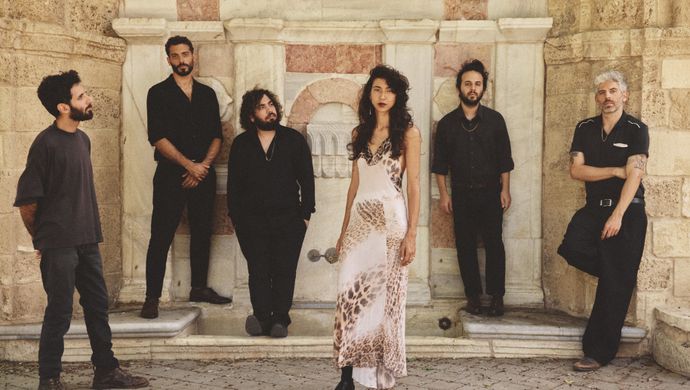 Zes bandleden staan bij een stenen muur. Eén persoon draagt een lichte jurk met patroon, de anderen zijn in donkere kleding gekleed.