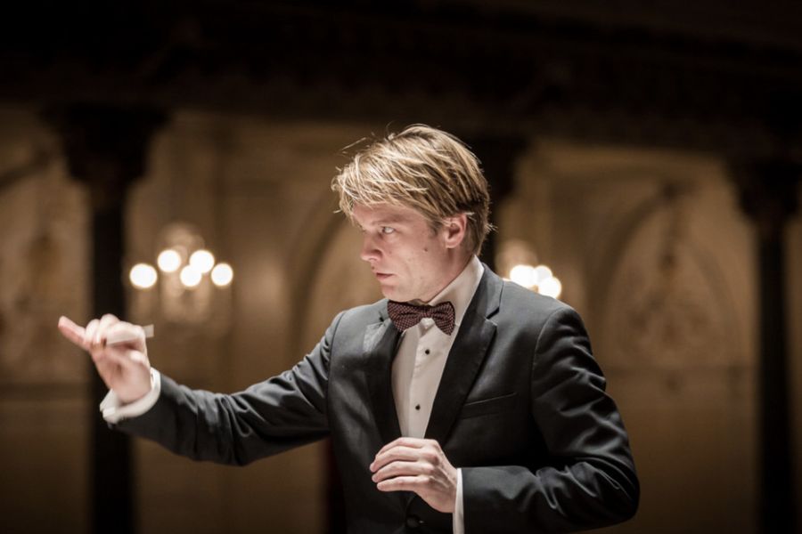 Dirigent in smoking met vlinderdas leidt een orkest in een klassieke concertzaal met warme verlichting en sierlijke muurschilderingen op de achtergrond.