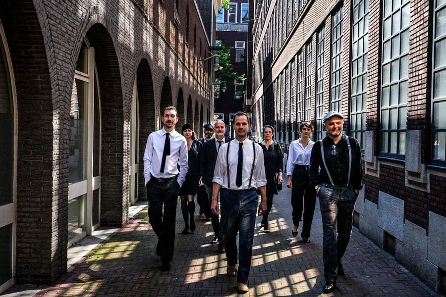 Group photo of six men dressed in dark suits with white shirts, standing in a straight line against a concrete wall. They look serious and professional toward the camera.