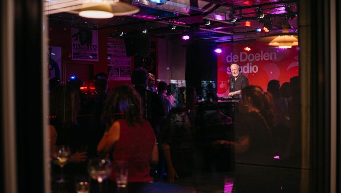 A cozy bar with purple and red lighting during the Disco Royale event. People are standing and sitting at tables while a dj stands on a small stage. The photo is taken from outside through a window.