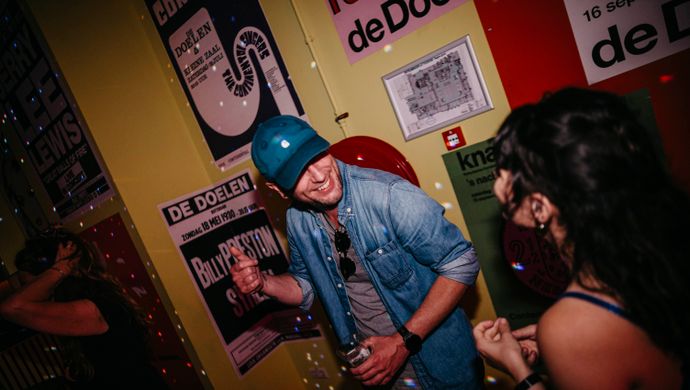 A man and a woman laugh and chat with drinks in hand during Disco Royale at de Doelen. They are standing by a brightly colored wall filled with retro posters, as disco ball lights move across the scene.