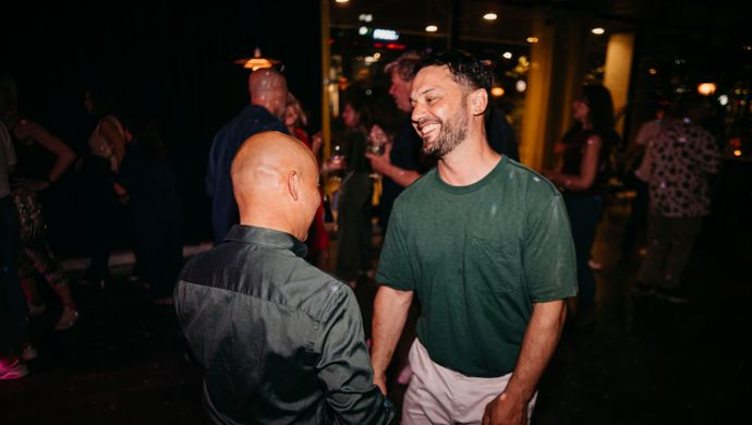 Two men are talking and laughing together during the Disco Royale event. They are in a warmly lit space with other people in the background also enjoying the evening.