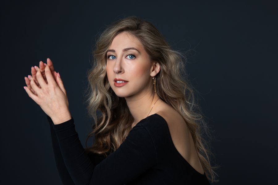 Vrouw met lang, golvend blond haar en elegante zwarte top poseert tegen een donkere achtergrond, met haar handen licht geheven en een rustige, open blik in de camera.