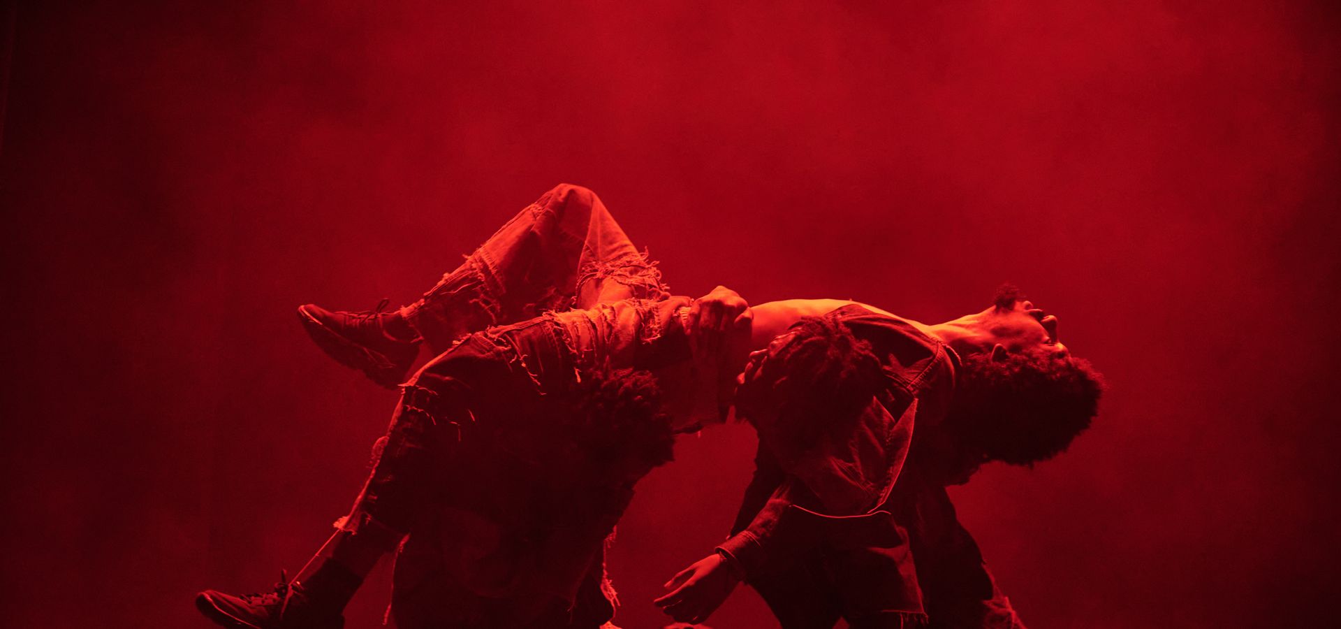 Twee dansers voeren een expressieve choreografie uit in rood toneellicht. Eén danser houdt de ander dramatisch gebogen over de schouders, terwijl rook of mist de achtergrond vult.