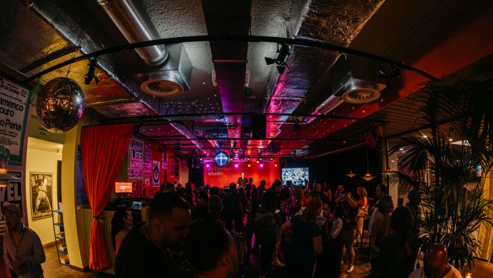 A crowded dance floor during Disco Royale at de Doelen Studio. The audience stands in a warmly lit space with red and purple disco lights, while a DJ performs on stage. Disco balls on the ceiling enhance the festive retro atmosphere.