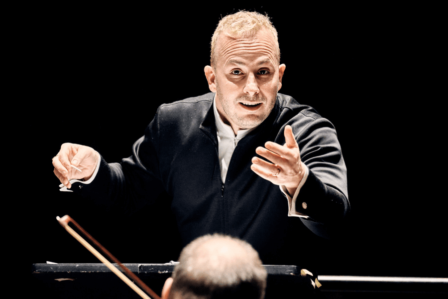 Een dirigent in zwarte concertkleding dirigeert intensief een orkest, met opgeheven handen en een geconcentreerde blik. De achtergrond is volledig zwart, waardoor de focus volledig op de dirigent ligt. Het hoofd van een muzikant is op de voorgrond zichtbaar.