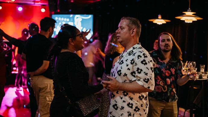 A couple dances together during Disco Royale at de Doelen Studio. They look at each other while other guests in the background are dancing or chatting. The room is warmly lit with red and golden tones, creating an intimate and festive atmosphere.