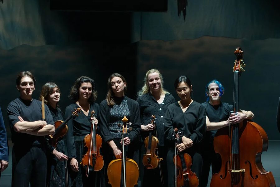Acht musici van een klassiek ensemble poseren in donkere kleding met hun strijkinstrumenten tegen een donkere, dramatische achtergrond. De groep bestaat uit violisten, altviolisten, cellisten en een contrabassist.