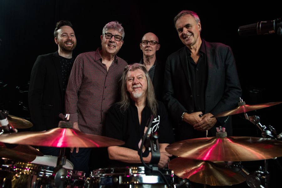 Vijf muzikanten poseren lachend achter een drumstel op een donker podium. De sfeer is informeel en warm, met focus op de drummer op de voorgrond.