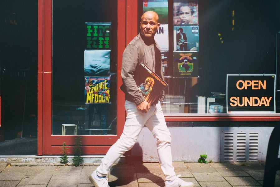Een man loopt lachend voorbij een winkel met rode kozijnen en een "OPEN SUNDAY"-bord in het raam. Hij draagt een beige jas, witte broek en witte sneakers, en kijkt over zijn schouder naar de camera.