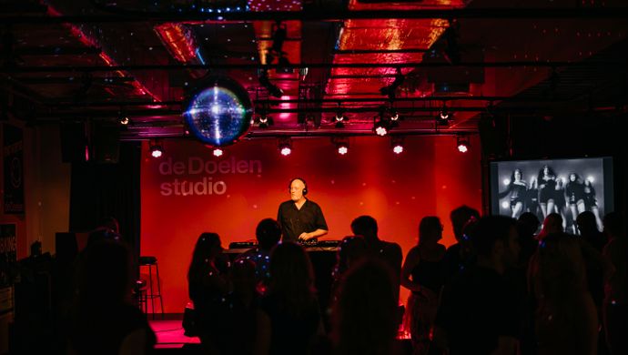 Audience watches a DJ performing on stage during Disco Royale at de Doelen Studio. The venue is lit in red and purple with a disco ball overhead and visuals displayed on a screen to the right. The atmosphere is energetic and festive.