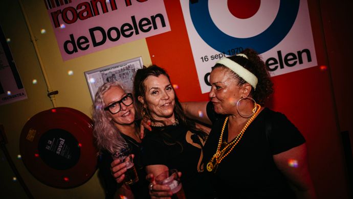 Three women pose together smiling during Disco Royale at de Doelen Studio. They stand close with drinks in hand, in front of a colorful wall filled with posters from de Doelen. 